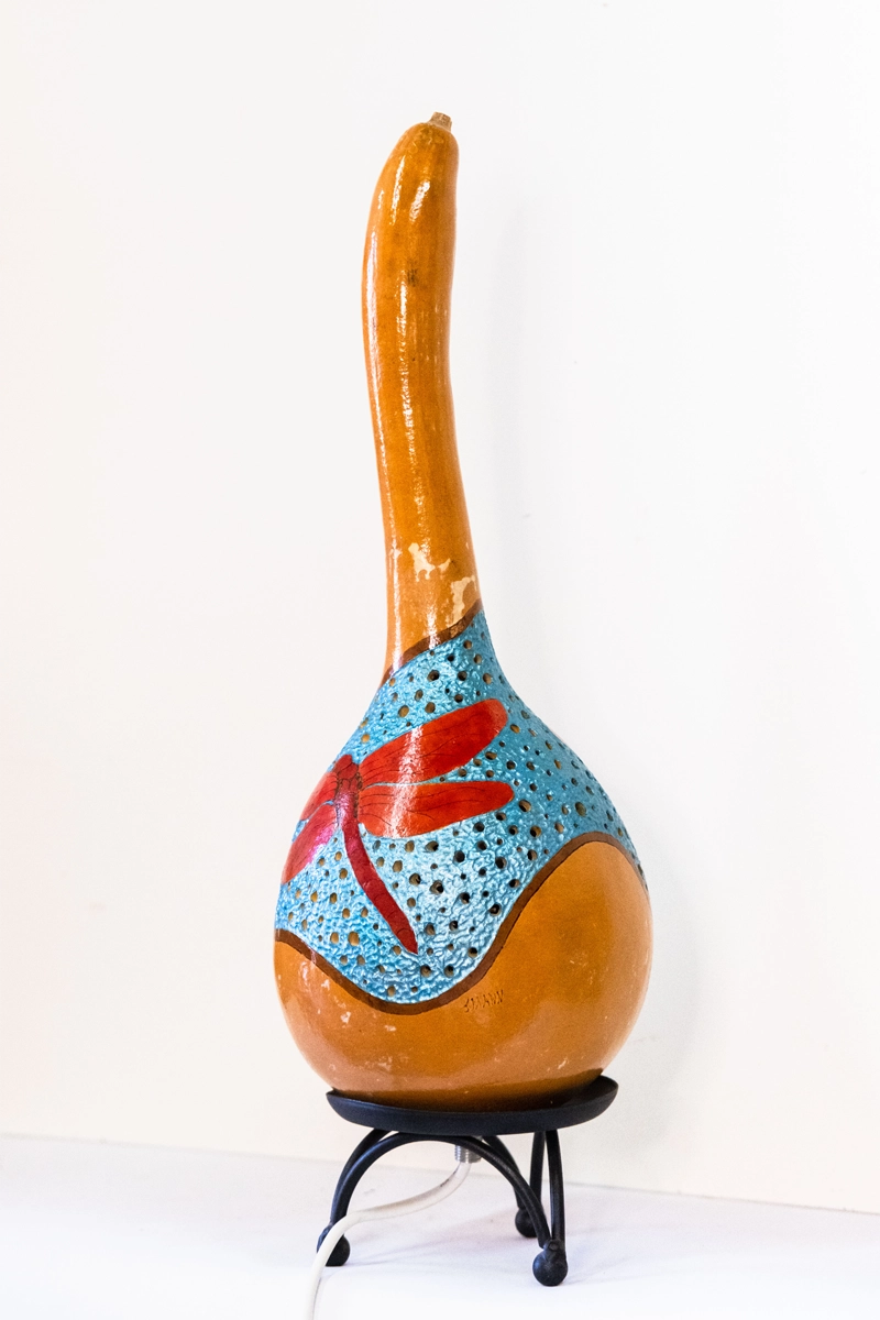 A gourd lamp depicting red dragonflies. Set on a black metal base. Wood-burnt, carved and finished in acrylics. Mains powered. 42cm tall.
