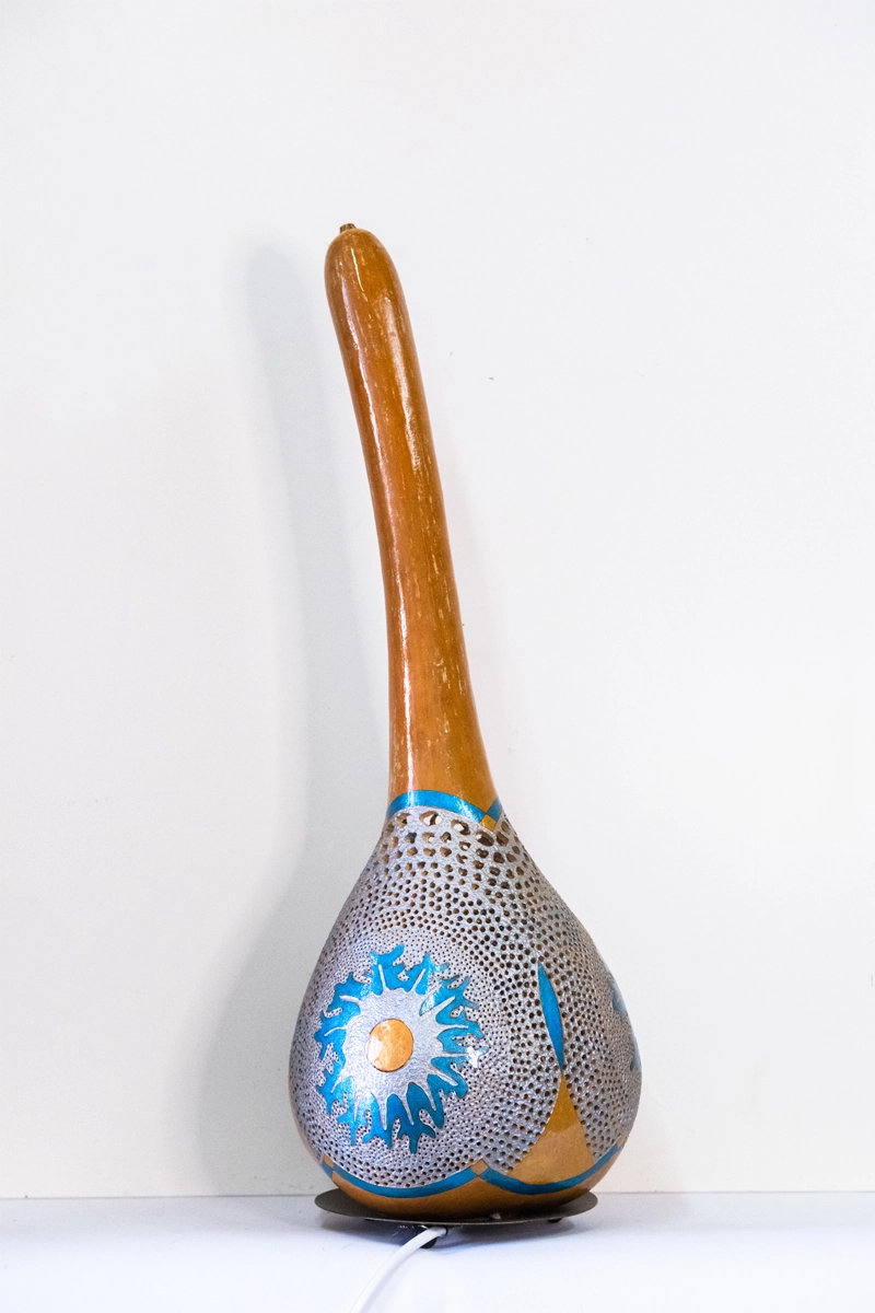 A gourd lamp with a delicately carved decreasing circular effect on three sides. Wood-burnt, carved and finished in acrylics on a circular metal base. Mains powered. 56cm tall