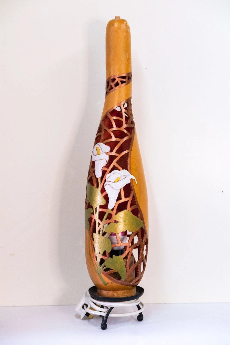 A delightfully carved gourd lamp, depicting Calla-lilies growing on trellis. Wood-burnt, carved and finished in wellburn inks, mounted on a metal tripod stand. Mains powered. 58cm tall.