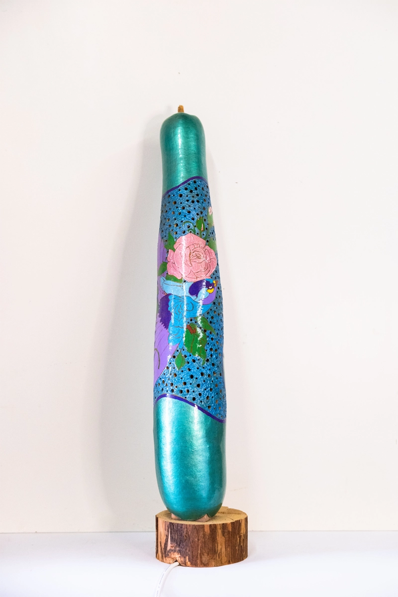 A gourd lamp depicting a bird and pink rose mounted on a hardwood base. Wood-burnt, carved and finished with acrylics. Mains powered. 60cm tall.