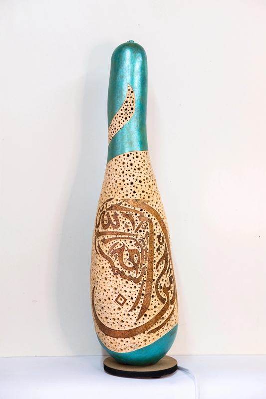 Gourd lamp wood-burnt and carved with Arabic lettering to create a female head . Finished in green metallic acrylic on a wooden base. Mains powered. 60cm tall.,