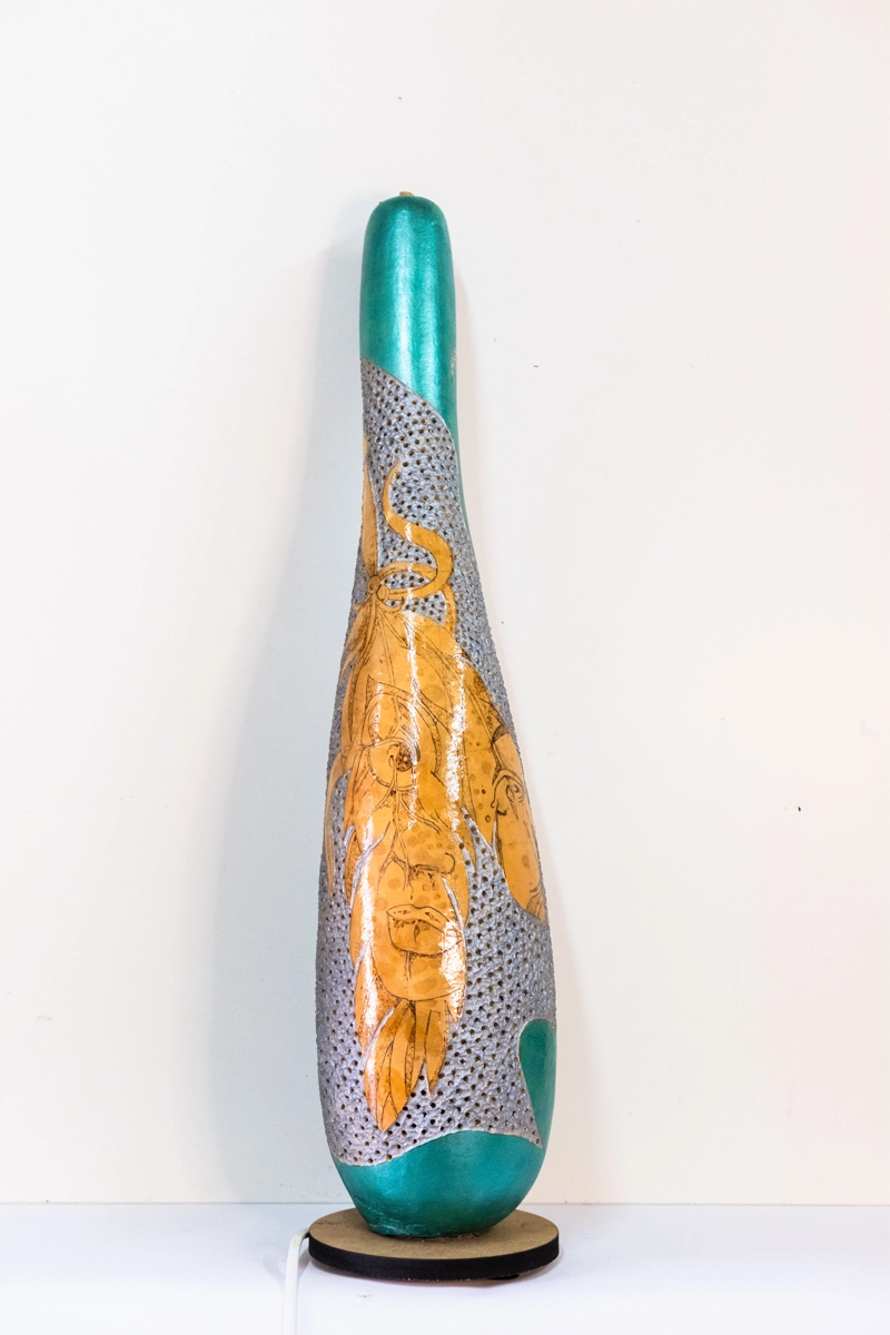 Gourd lamp wood-burnt and carved to form a face on a feather, finished with acrylics on a wooden base. Mains powered. 58cm tall